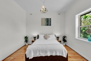 A bedroom with a wooden floor, a bed with white bedding, two bedside tables with lamps, a chandelier, a window with a view of greenery, and a painting on the wall. - Charming Family Retreat with Arcade + Walk to Shops - Glenunga (Glenunga)