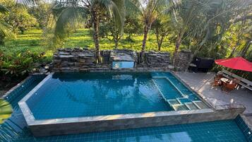 Una piscina con paredes de piedra, rodeada de vegetación y un patio con una sombrilla roja.