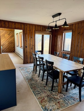 Een eethoek met een houten tafel en stoelen, een kamerbreed tapijt met een patroon, en uitzicht op de natuur door grote ramen.
