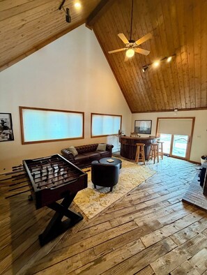 A loft-style room with a wooden ceiling, a foosball table, a leather sofa, and a bar area.