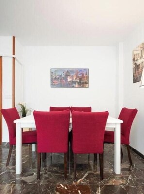 A dining area with a white table and red chairs, a marble floor, and a cityscape painting on the wall.