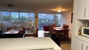 A modern kitchen with white cabinets, a dining area with wooden chairs, a sofa, and a large window offering a city view. - Apartment in San Pedro Super Central and Safe Zone (San Pedro Garza García)