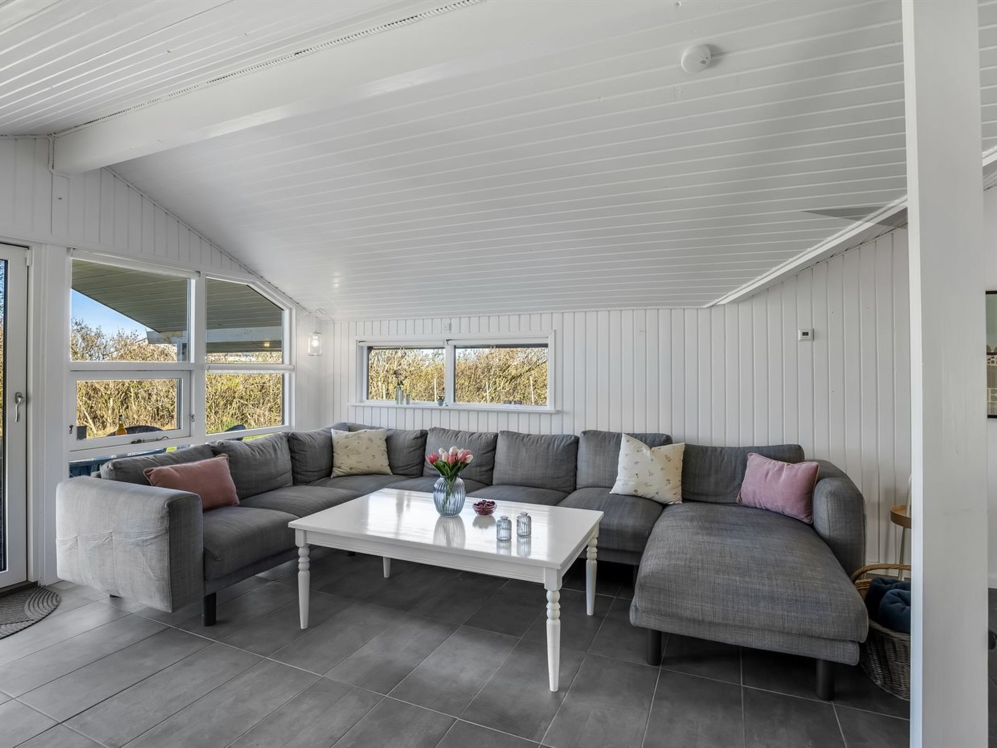 Un salon moderne avec un canapé d’angle, une table basse blanche et de grandes fenêtres donnant sur la verdure.