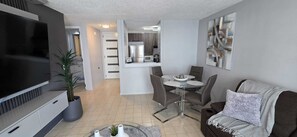 Ein modernes Wohnzimmer mit Flachbildfernseher, einer Couch, einem Esstisch mit Stühlen, einer Küchenzeile inklusive Kühlschrank und Mikrowelle sowie einem Flur, der in einen weiteren Raum führt.