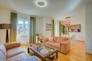 A living room with a sofa, a coffee table, a dining area with a table and chairs, a mirror, and a view of the outside. - Casa Berna - Balcony Apt with Mountain Views, Lugano, Switzerland (Lugano)