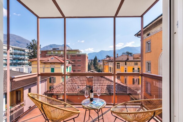Uma varanda com cadeiras de vime e uma mesa, com vista para um horizonte urbano com edifícios e montanhas.