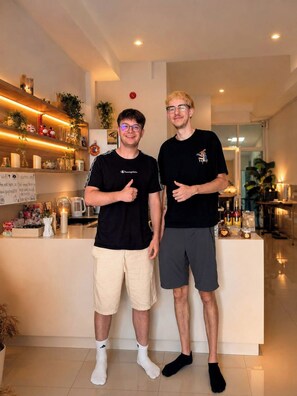 Two individuals standing in a modern interior space with a kitchen counter, shelves with various items, and a visible clock on the wall. - Behome and hostel (Chiang Mai)