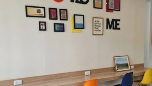 Uma parede com quadros e um disco de vinil, três cadeiras em tons de rosa, laranja e azul, e uma prateleira de madeira.