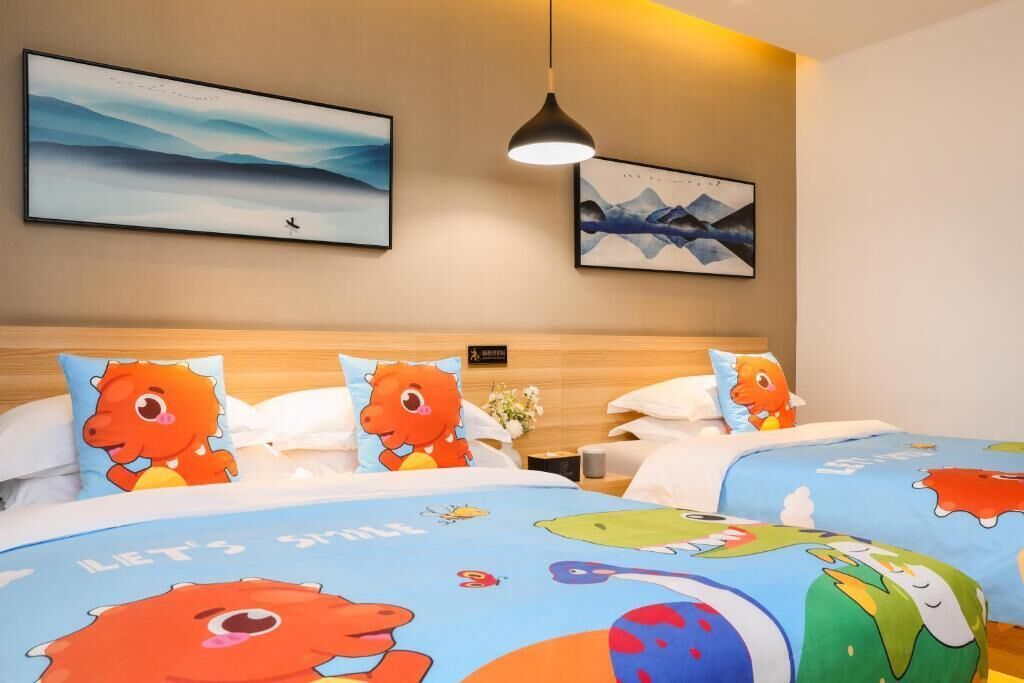 A hotel room with two beds, each with a colorful bedspread featuring cartoon characters. There are two framed artworks on the wall above the beds.