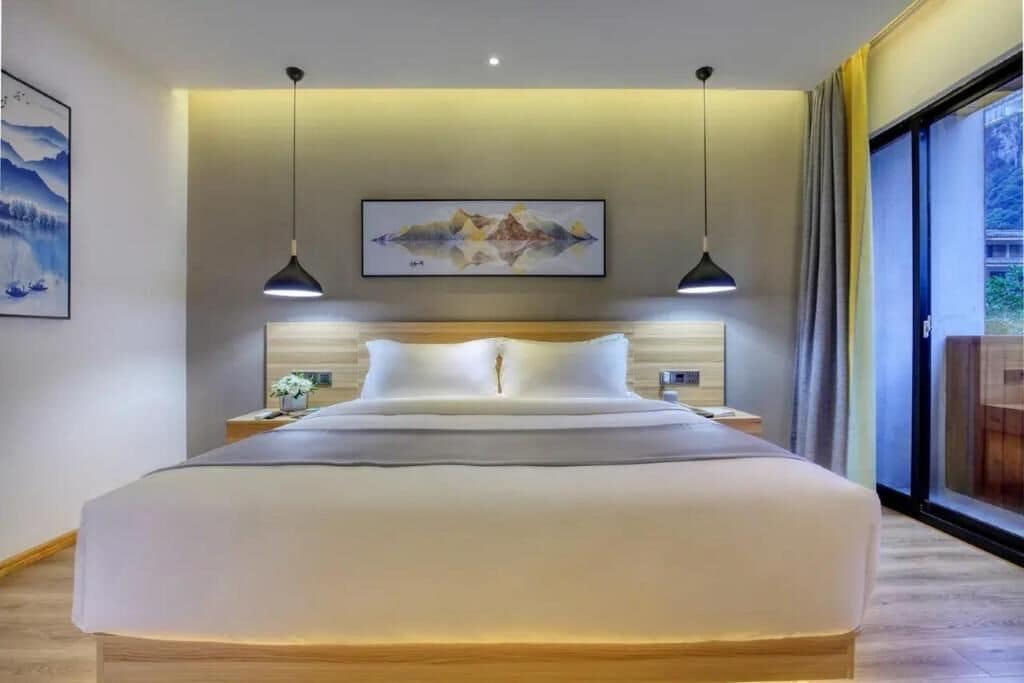 Un dormitorio de hotel moderno con una cama grande, dos lámparas colgantes, un cuadro en la pared y una ventana con cortinas.