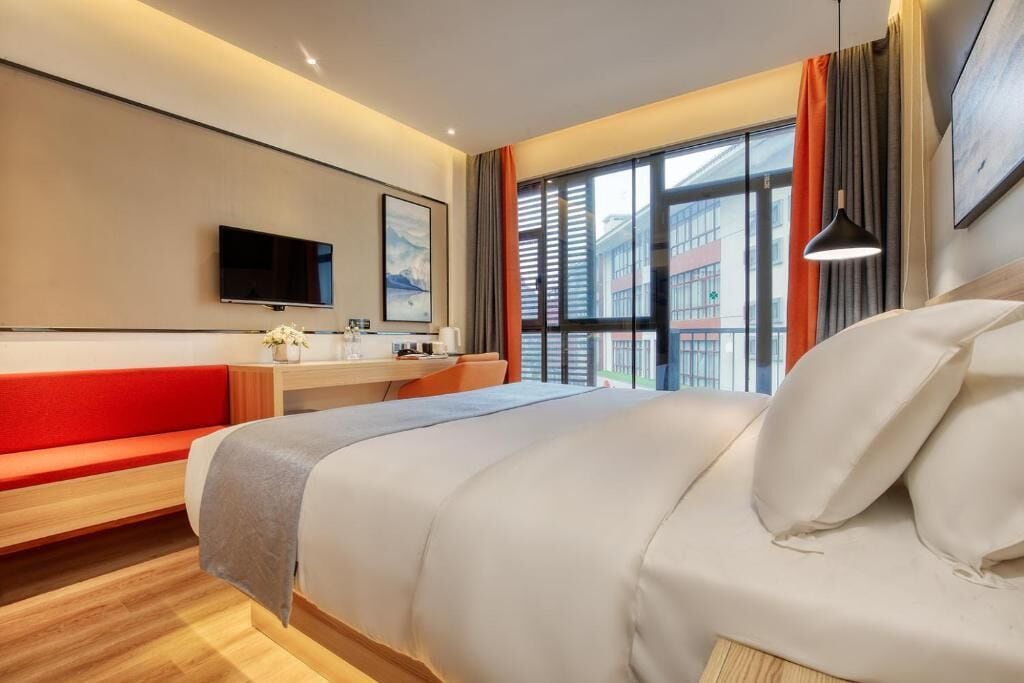 A modern hotel room with a bed, a desk, a TV, and a window with blinds.