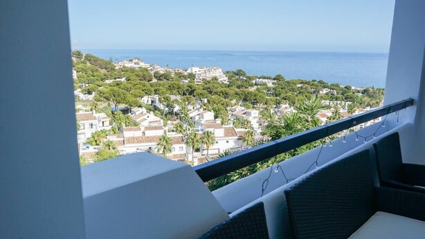 Un balcón con vistas a un pueblo costero, paredes blancas y una silla de mimbre.