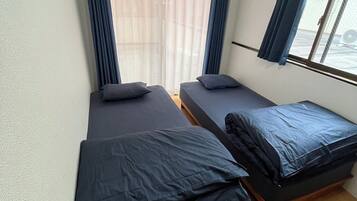 A room with two single beds, a window with curtains, an air conditioning unit, and a wooden floor.