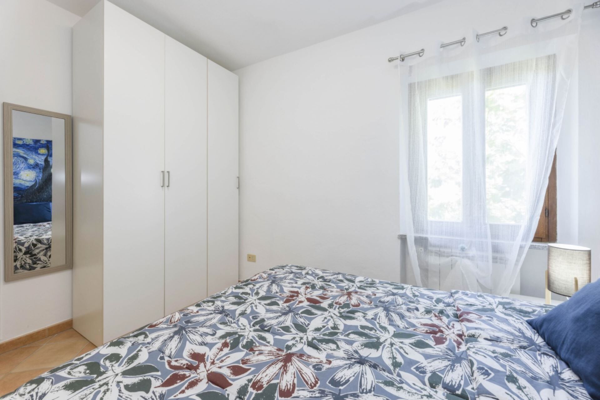 Ein Schlafzimmer mit Bett, Schrank, einem Bild an der Wand und einem Fenster mit Vorhängen.