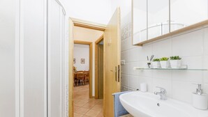 Een badkamer met een wit wastafelmeubel, een glazen plank met planten en een houten deur die naar een eetruimte leidt.