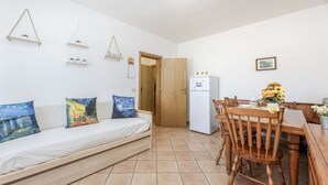 Een compacte woonruimte met een bank, een houten eettafel, stoelen, een koelkast en een deur naar een andere kamer.