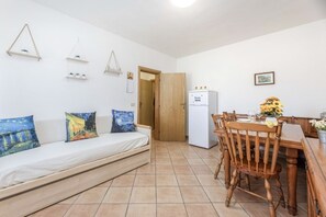A compact living space with a sofa, wooden dining table, chairs, a refrigerator, and a door leading to another room. - Bilocale Vincent - Goelba (Campo nell'Elba)