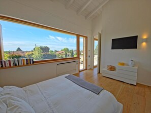 Ein Schlafzimmer mit großem Fenster, einem Bett, einem Fernseher und einer kleinen, weißen Kommode.