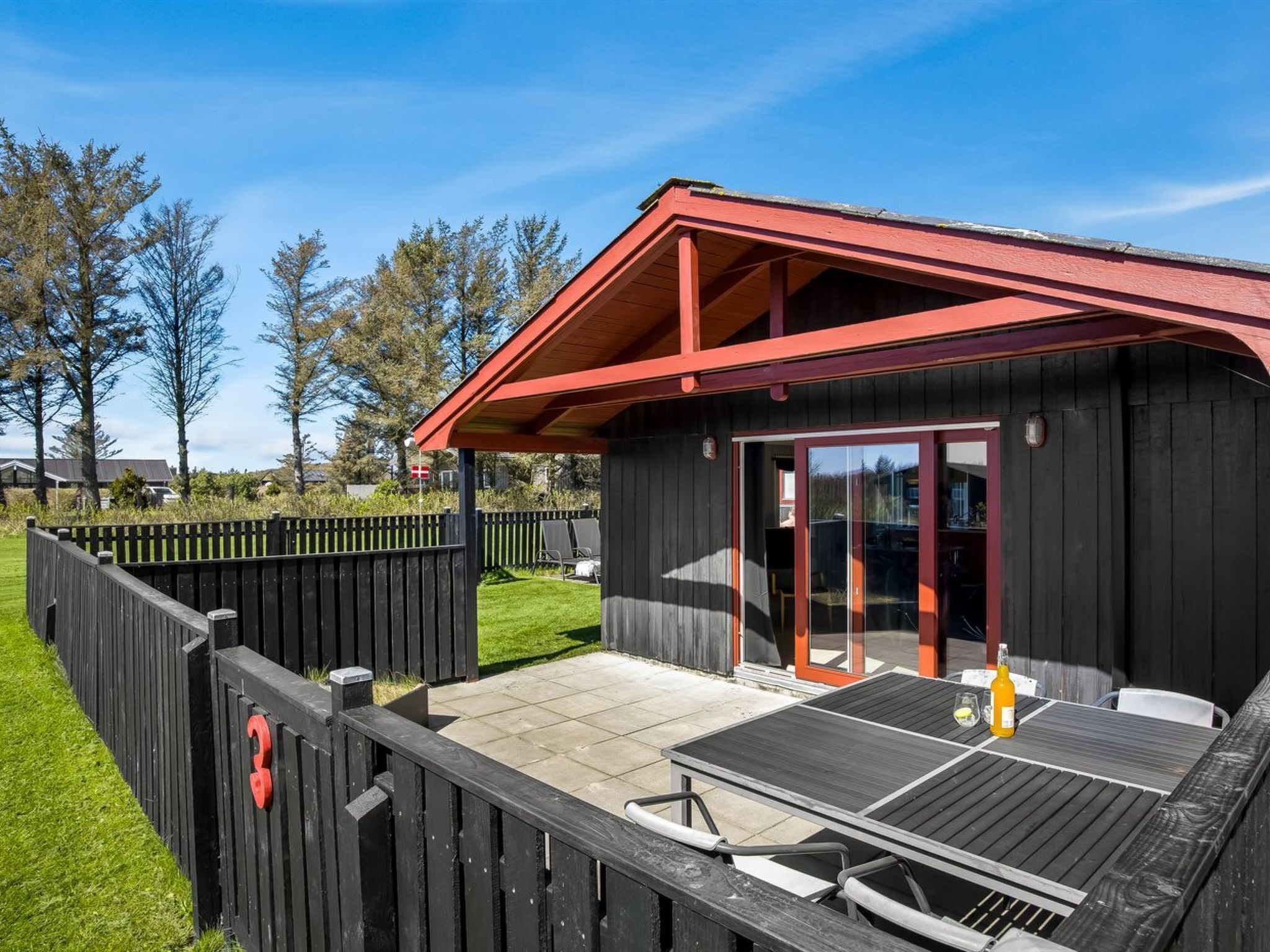 Un bâtiment en bois avec un toit rouge, une terrasse avec une table et des chaises, et un espace clôturé.
