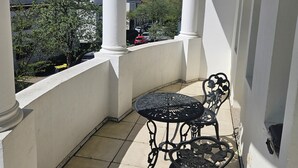 En balkon med hvide søjler, et lille bord med en stol, samt udsigt til bygninger og træer.