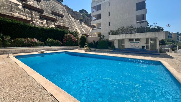 A swimming pool with a clear blue water, surrounded by a paved area and a building with multiple floors and balconies.