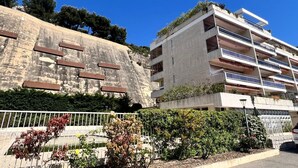 A multi-story building with balconies, a stone wall with metal bars, and greenery.