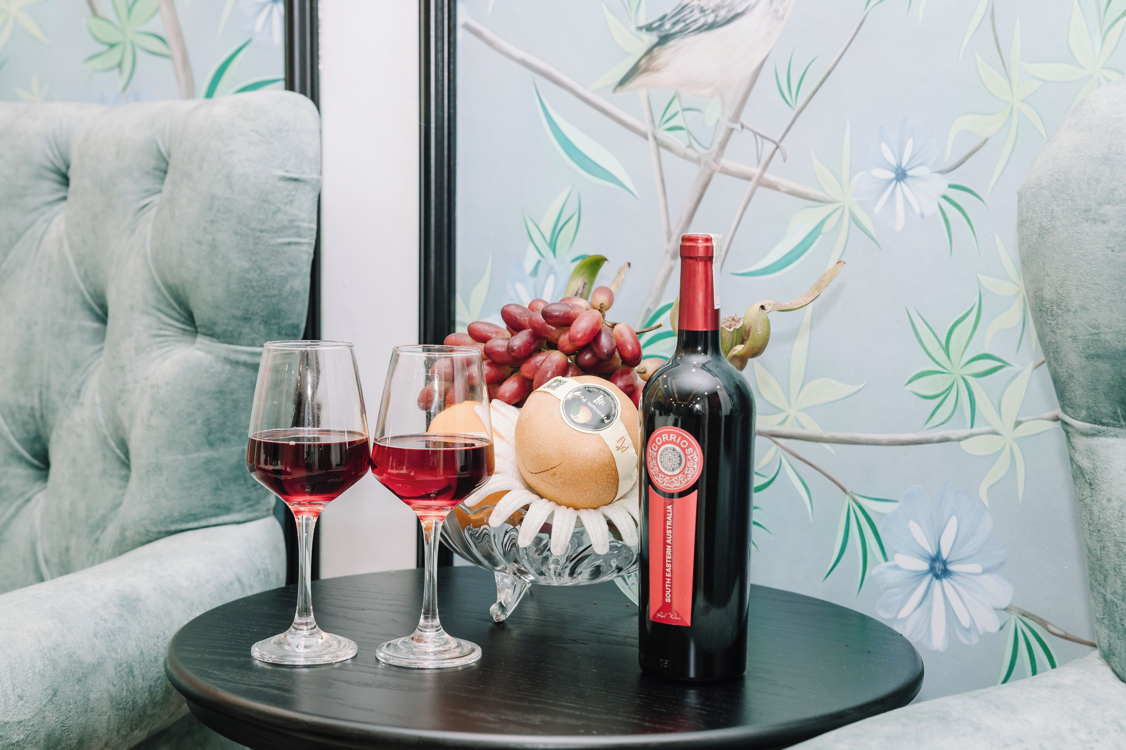 A table with two wine glasses, a bottle of wine, a decorative fruit bowl, and a plush armchair.