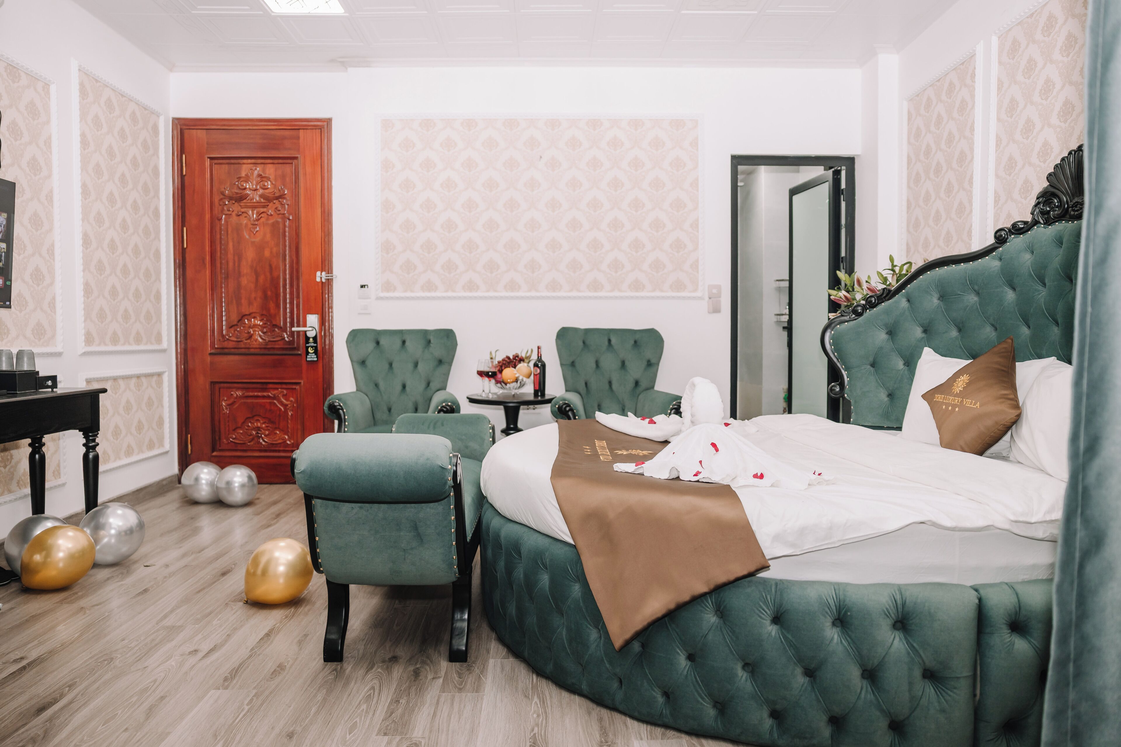 A hotel room with a green upholstered bed, a red door, a small table with a vase, and a green armchair.