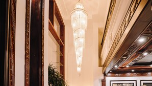 Um corredor imponente com um lustre grande e ricamente decorado, painéis de madeira e uma escadaria com molduras detalhadas.