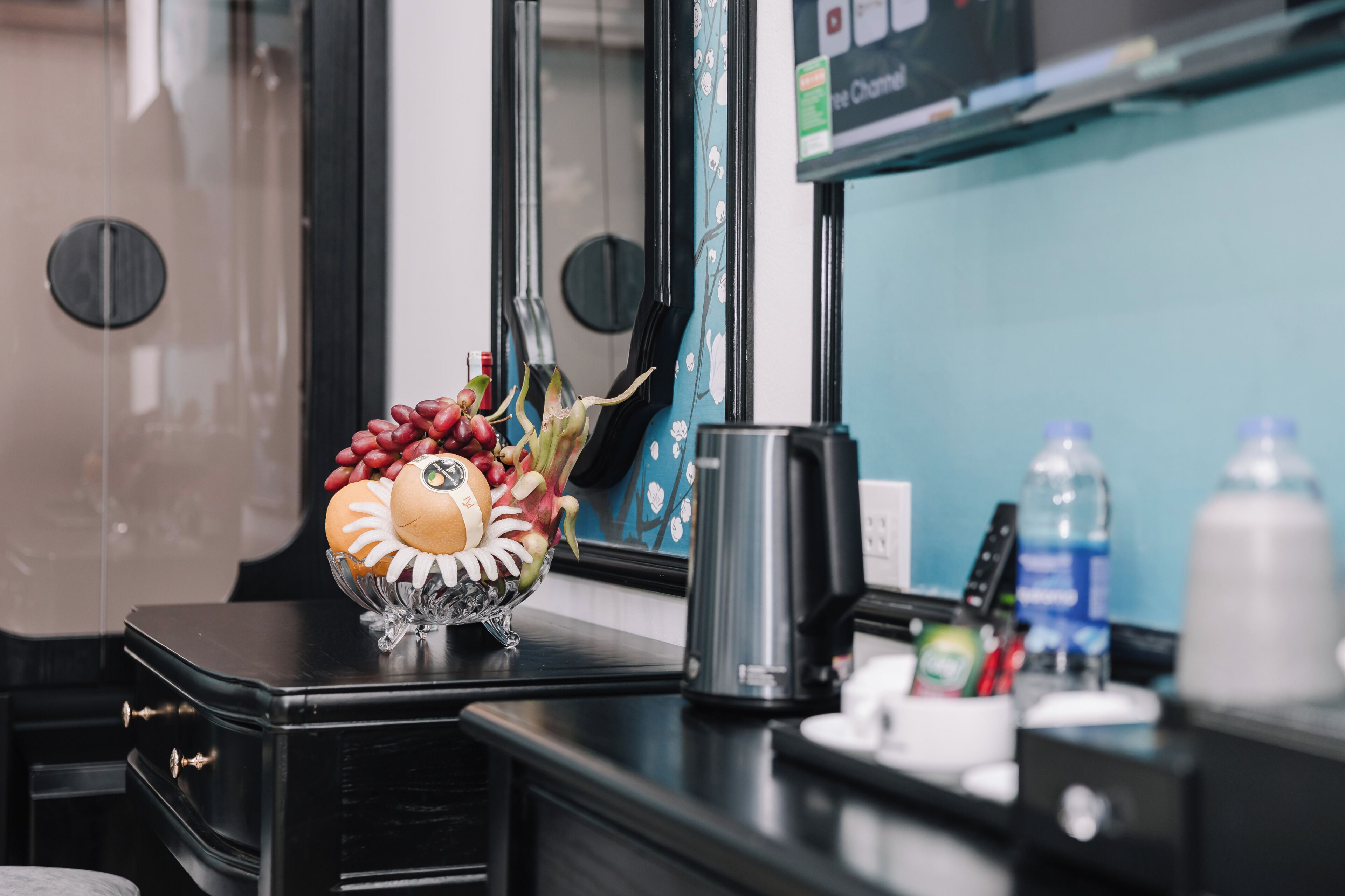 Habitación de hotel con un escritorio negro, un cuenco decorativo con frutas, una cafetera y una pantalla de televisión.