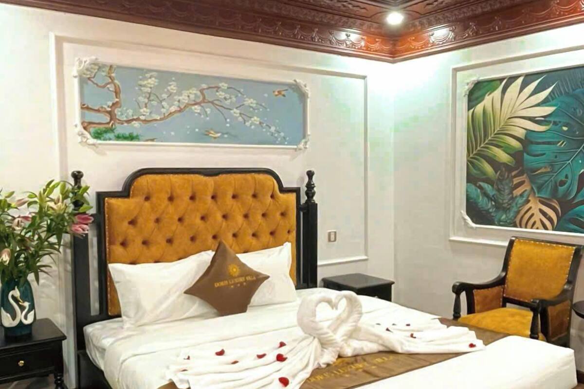 Ein stilvoll eingerichtetes Schlafzimmer mit einer gepolsterten Kopfteile, einem Bett mit weißen Leinen, einem dekorativen Schwanenkissen und einem Nachttisch mit Blumen.