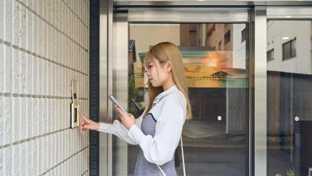 A person standing at a door, holding a phone, and interacting with a doorbell. - Nemuru Kagoshima-Chuo Station (Kagoshima)