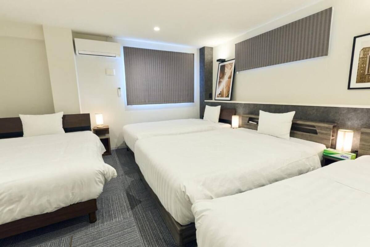 Ein Hotelzimmer mit drei Betten, einem großen Fenster mit Jalousien, einem gerahmten Bild an der Wand und einem kleinen Tisch mit Lampe.