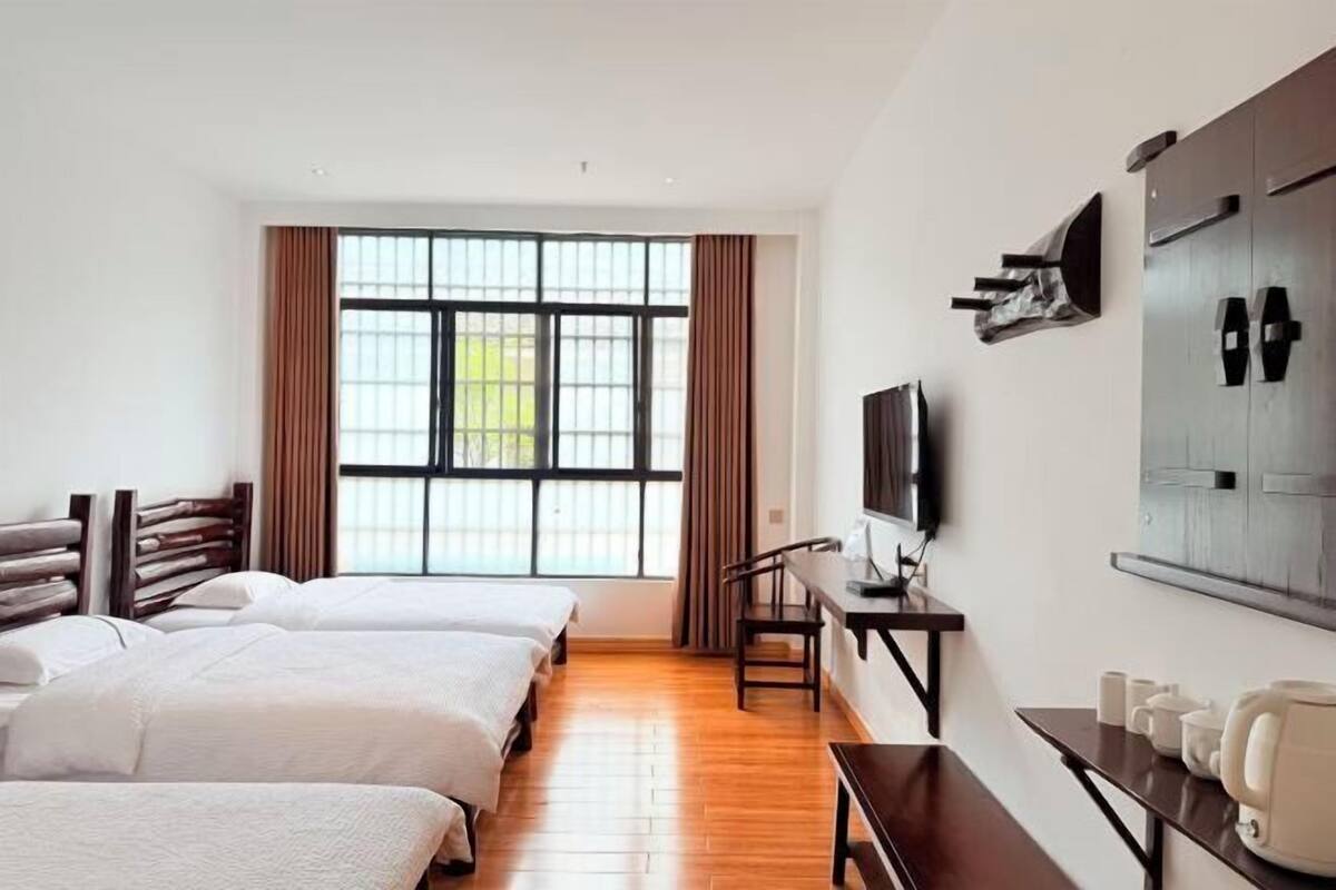 Une chambre d’hôtel avec trois lits, une télévision, un bureau avec une chaise et une grande fenêtre avec des rideaux.