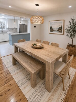 A dining area with a wooden table, benches, wicker chairs, a hanging light fixture, a framed landscape painting, and a potted plant. - Golden Slumbers, your coastal retreat in Goose Rocks Beach, Kennebunkport (Kennebunkport)