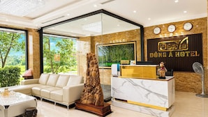 A hotel reception area with a marble reception desk, a white sofa, a decorative wooden sculpture, and a view of greenery through large windows. - Dong A Hotel Pho Co Ninh Binh (Hoa Lu)