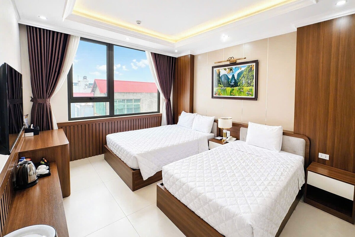 A hotel room with two beds, a desk with a kettle, a TV, and a painting on the wall.