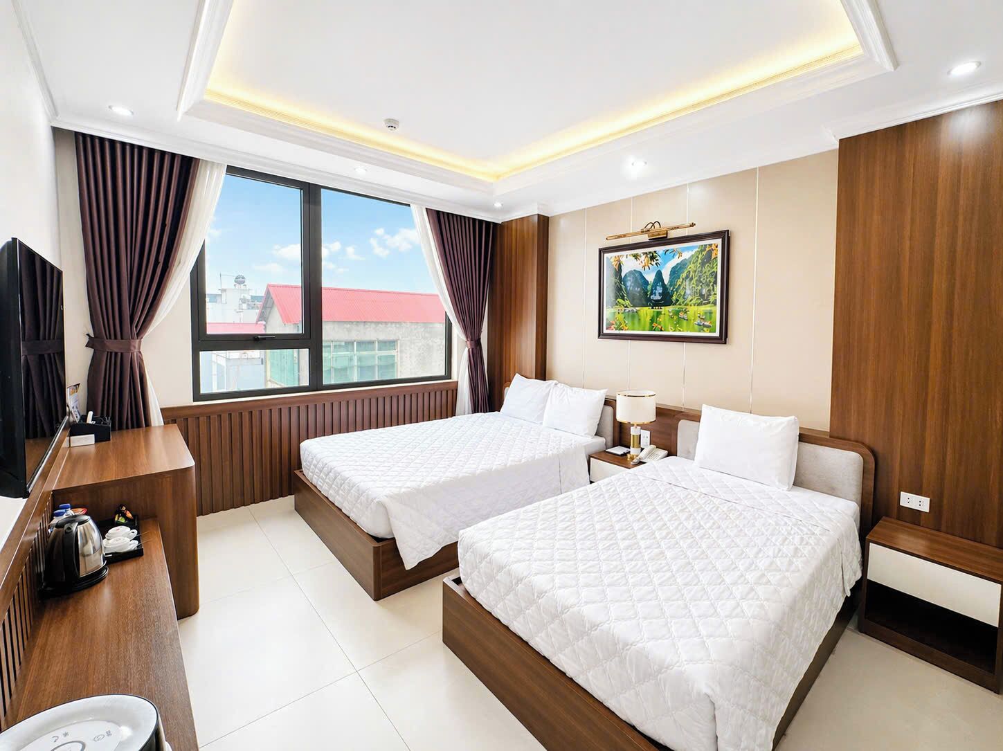 A hotel room with two beds, a desk with a kettle, a TV, and a painting on the wall.