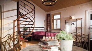 Una habitación acogedora con una escalera espiral, una cama con una colcha roja, una mesa de madera con libros y una planta, y una ventana con cortinas.