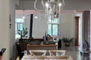 Zona pranzo con lampadario, sedie in legno, isola centrale con utensili da cucina e ampie finestre.