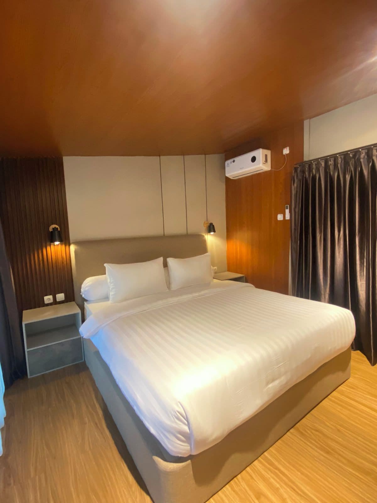 A hotel room with a large bed, bedside tables, a wall-mounted air conditioner, and a curtain.