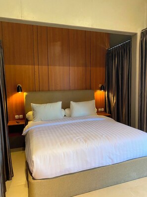 A hotel room with a large bed, wooden panel headboard, two bedside lamps, and a curtain. - The Foresta By Ohana (Yogyakarta)