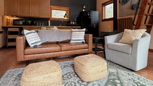 A cozy living room with a brown leather sofa, a grey armchair, and two woven ottomans. A kitchen with wooden cabinets and a black refrigerator is visible in the background.