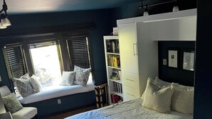 A bedroom with a bed, a window seat, a bookshelf, and a desk.