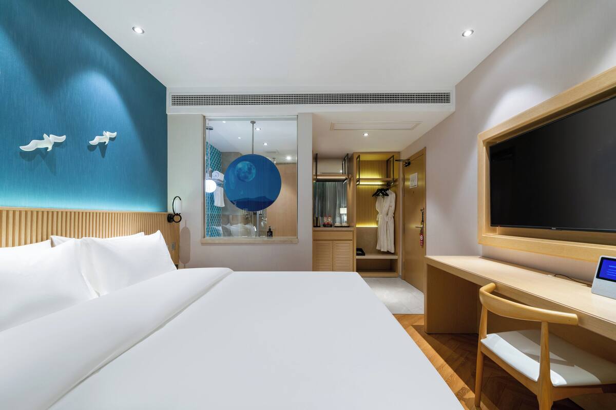 A modern hotel room with a large bed, a desk with a computer, a TV, and a walk-in closet.