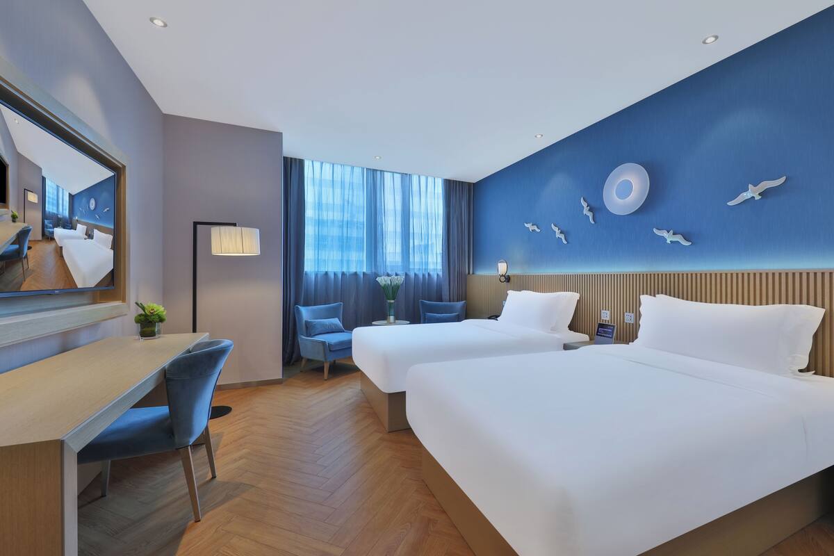 A hotel room with two beds, a desk, chairs, a large mirror, and a view of the city.