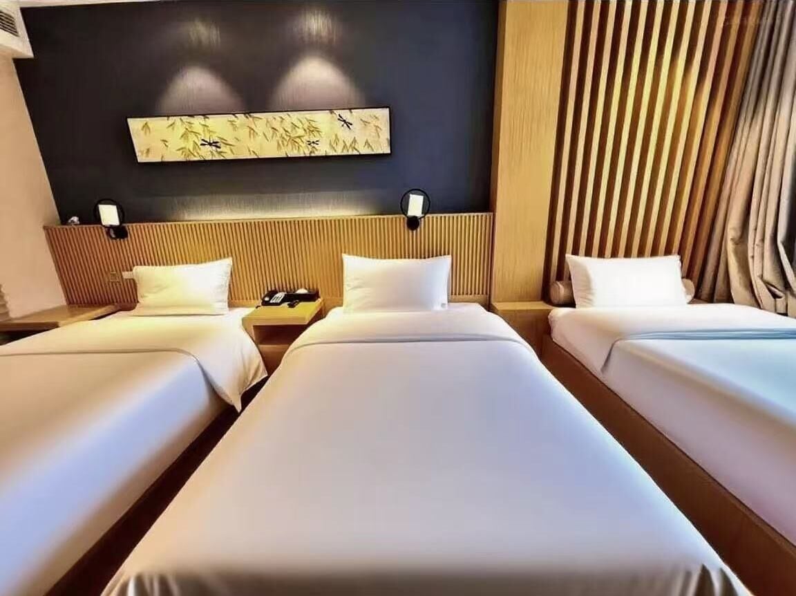 Een hotelkamer met twee eenpersoonsbedden, een houten hoofdbord, een schilderij aan de muur en een raam met verticale jaloezieën.