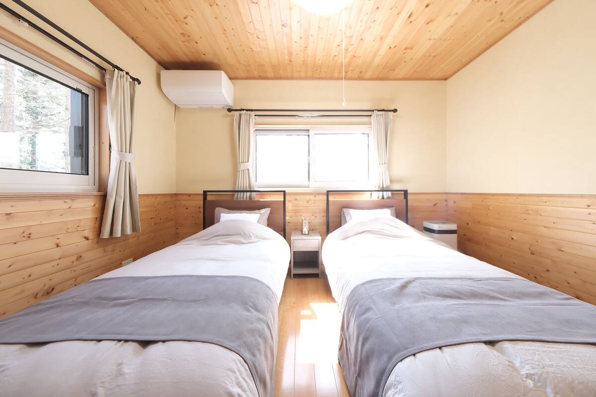 A room with two beds, wooden walls, a window with curtains, an air conditioning unit, and a nightstand with a lamp.