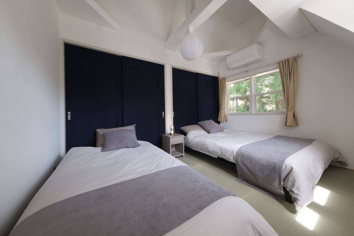 A room with two beds, a window with curtains, a nightstand, and a ceiling fan.