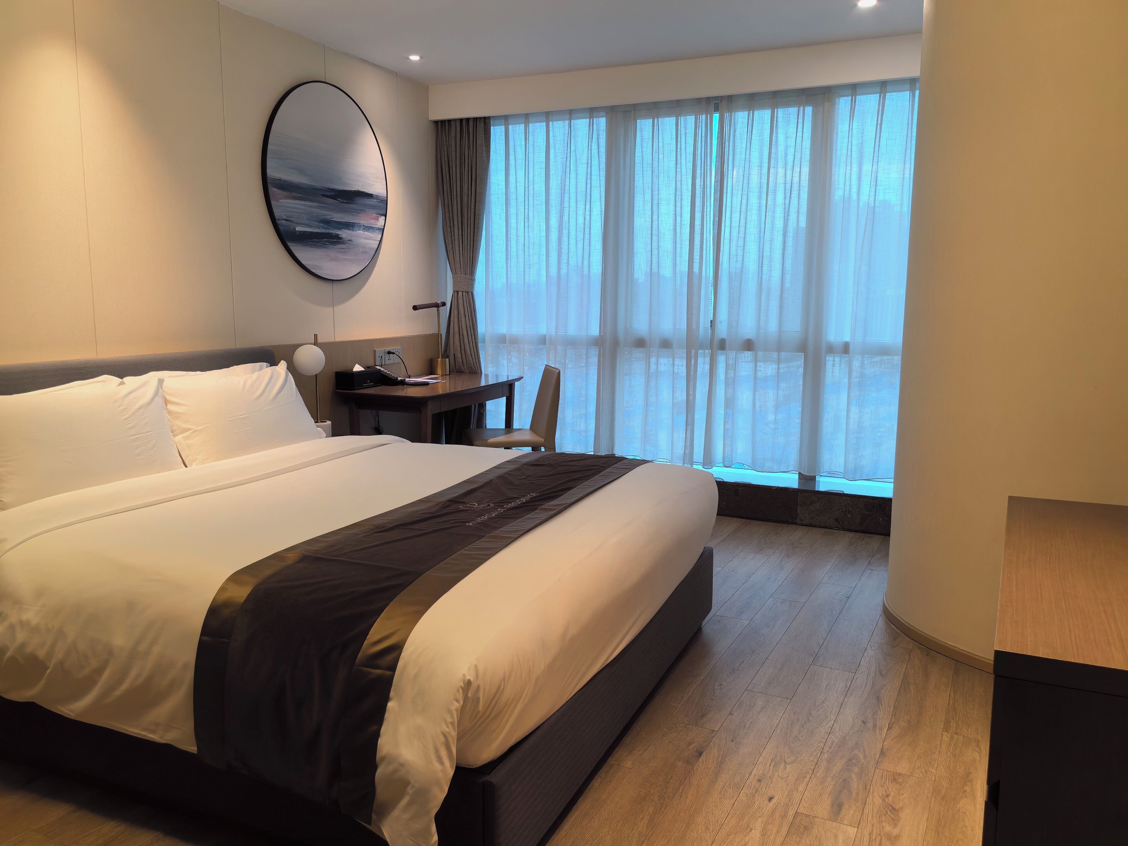 A hotel room with a large bed, a desk, a chair, and a large window with curtains.